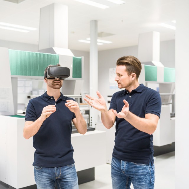 Zwei junge Männer, von denen einer eine Virtual-Reality-Brille trägt 