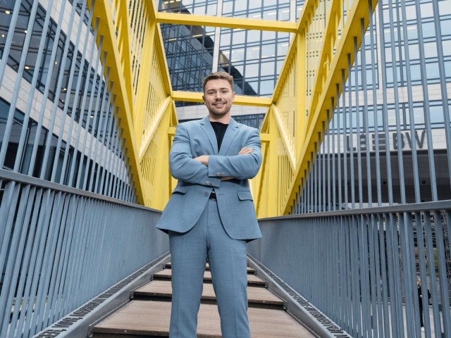 Ein Mann steht mit verschr&auml;nkten Armen auf einer Br&uuml;cke mit gelbem Gel&auml;nder.