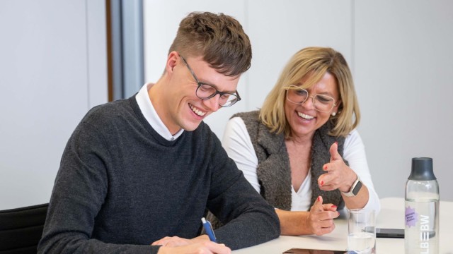 2 Personen sitzen an einem Tisch und arbeiten zusammen, wobei die ältere der jüngeren Person etwas erklärt.
