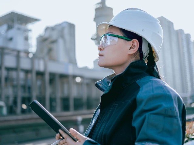 Eine Frau mit Helm, Schutzbrille und Tablet beobachtet Bauarbeiten