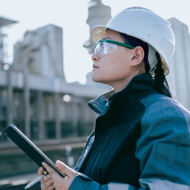Eine Frau mit Helm, Schutzbrille und Tablet beobachtet Bauarbeiten