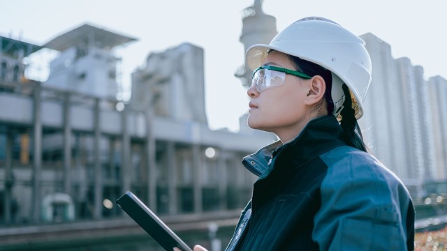 Eine Frau mit Helm, Schutzbrille und Tablet beobachtet Bauarbeiten