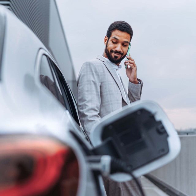 Mann lehnt an einem Elektroauto während es an eine Ladestation angeschlossen ist.