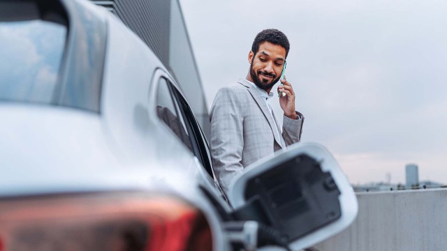 Mann lehnt an einem Elektroauto während es an eine Ladestation angeschlossen ist.