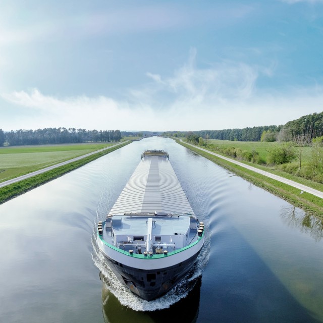 Frachtschiff fährt auf einem Fluss in Frontalansicht, im Hintergrund Natur und blauer Himmel
