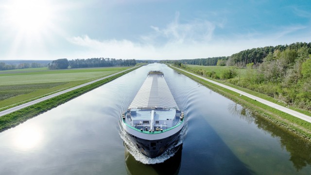 Frachtschiff fährt auf einem Fluss in Frontalansicht, im Hintergrund Natur und blauer Himmel