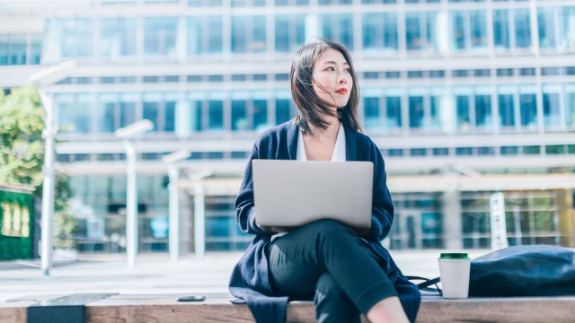 Frau sitzt auf einer Bank mit Laptop auf dem Schoß und einem Kaffeebecher neben ihr