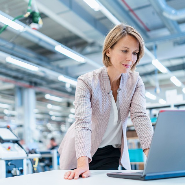 Geschäftsfrau steht in moderner Fabrikhalle am Laptop