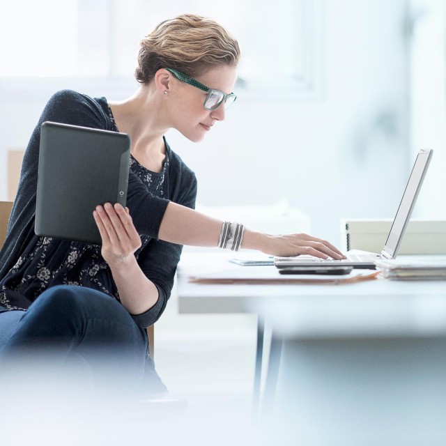 Blonde Frau mit grüner Brille arbeitet am Schreibtisch und hält Tablet in einer Hand