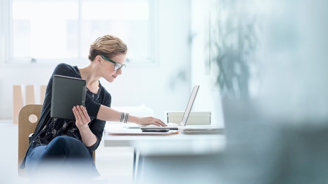 Blonde Frau mit grüner Brille arbeitet am Schreibtisch und hält Tablet in einer Hand