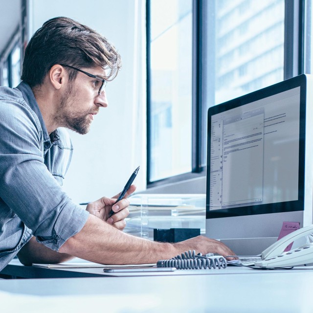 Mann mit Brille und hellblauem Hemd sitzt arbeitend am Schreibtisch und blickt auf einen Monitor