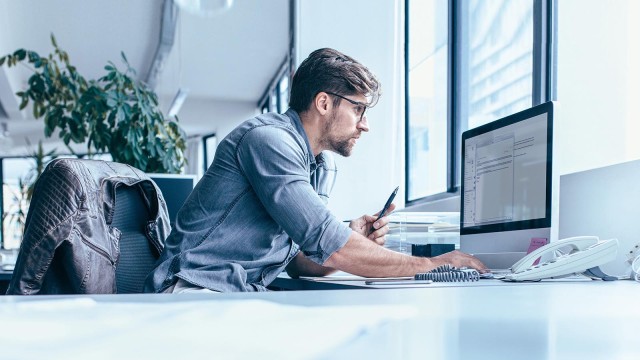Mann mit Brille und hellblauem Hemd sitzt arbeitend am Schreibtisch und blickt auf einen Monitor